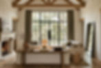 Organic modern living room with double-height ceilings, raw plaster walls, exposed oak beams, a large black steel grid window with linen drapes overlooking trees, a rustic reclaimed wood console table styled with a terracotta jug, stacked books, and a brass lamp, with wide plank oak floors and a woven rattan basket.