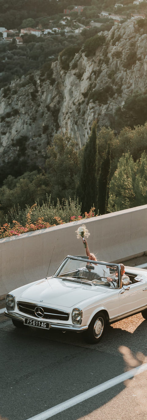 Sur une route en corniche, un couple de mariés dans un cabriolet blanc et la mariée lève son bouquet de fleurs - voiture de mariage - photographe de mariage Cannes, Monaco, Côte d'Azur