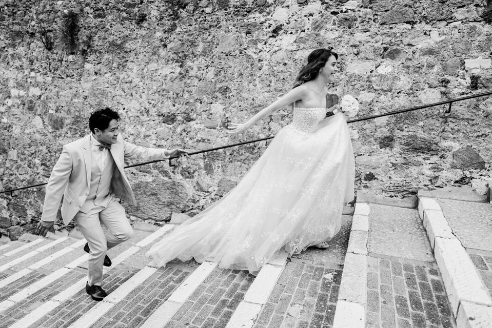 Dans des escaliers de vieille pierre, un jeune marié court en riant après sa femme qui a pris de l'avance - noir et blanc - photographe de mar