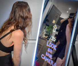 A woman with wavy brown hair leans towards an interactive mirror photo booth, which displays the prompt "LOOK at the CAMERA" with colorful graphics. The engaging display and instructions highlight the interactive and fun experience provided by our photo booth, perfect for capturing memorable moments at any event.