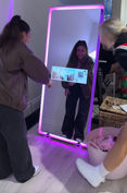 A young woman interacts with a full-length mirror photo booth, framed with vibrant pink neon lights. She smiles as she holds a photo strip showing her captured moments, highlighting the fun and engaging experience. This image illustrates the excitement and novelty of using our modern photo booth, perfect for any event to create memorable keepsakes.