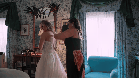 Two women in a floral room, one helping the other in a white dress get ready by a mirror, with a blue chair and bright window.
