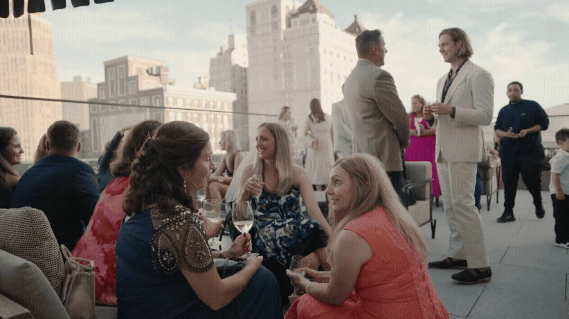 People socializing at a rooftop gathering during a wedding cocktail hour at Book Tower in Detroit, Michigan. Women in bright dresses sit talking and laughing, while others stand chatting. Cityscape in the background.