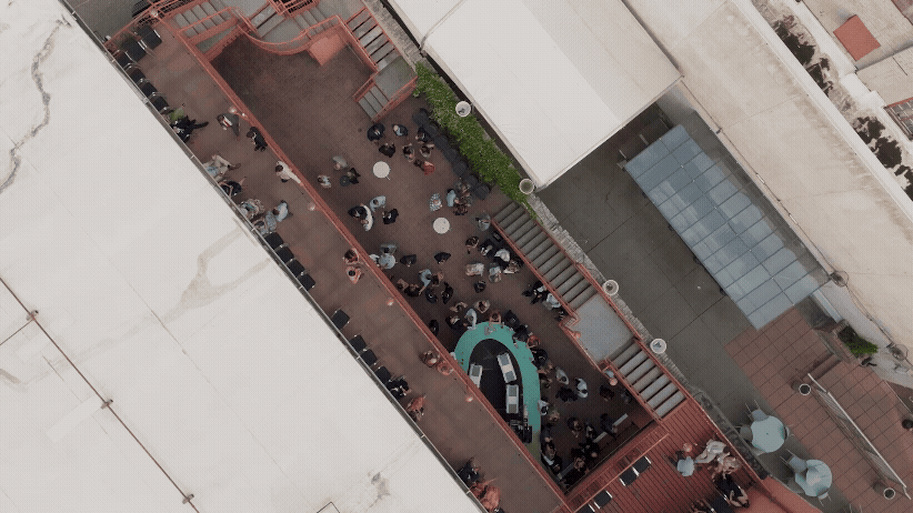 Aerial view of an alley with people gathered around a curved green bar at Crofoot in Pontiac Michigan during a wedding cocktail hour. The setting is urban with red-tiled flooring and white canopies.