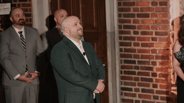 Groom in green suit smiling at the bride walking down the steps at the Detroit Institute of Arts in Michigan. Two groomsmen in suits stand behind him, observing. Warm lighting, formal setting.