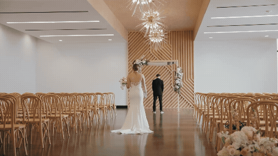 Bride in white gown and groom walk down a modern wedding aisle with wooden chairs, warm wood slats, and hanging lights.