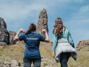 Two people facing the Old Man of Storr rock pile on the Isle of Skye, one is pointing to the back of their shirt to the Macbackpackers logo.