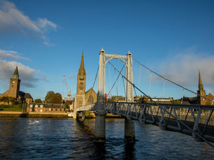A metal bridge crossing a river with three church spires and the Inverness city skyline in the background.