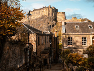 Edinburgh Castle as viewed from the Vennel Steps with the roofs of houses in the foreground, a lamppost, and a tree with orange leaves.