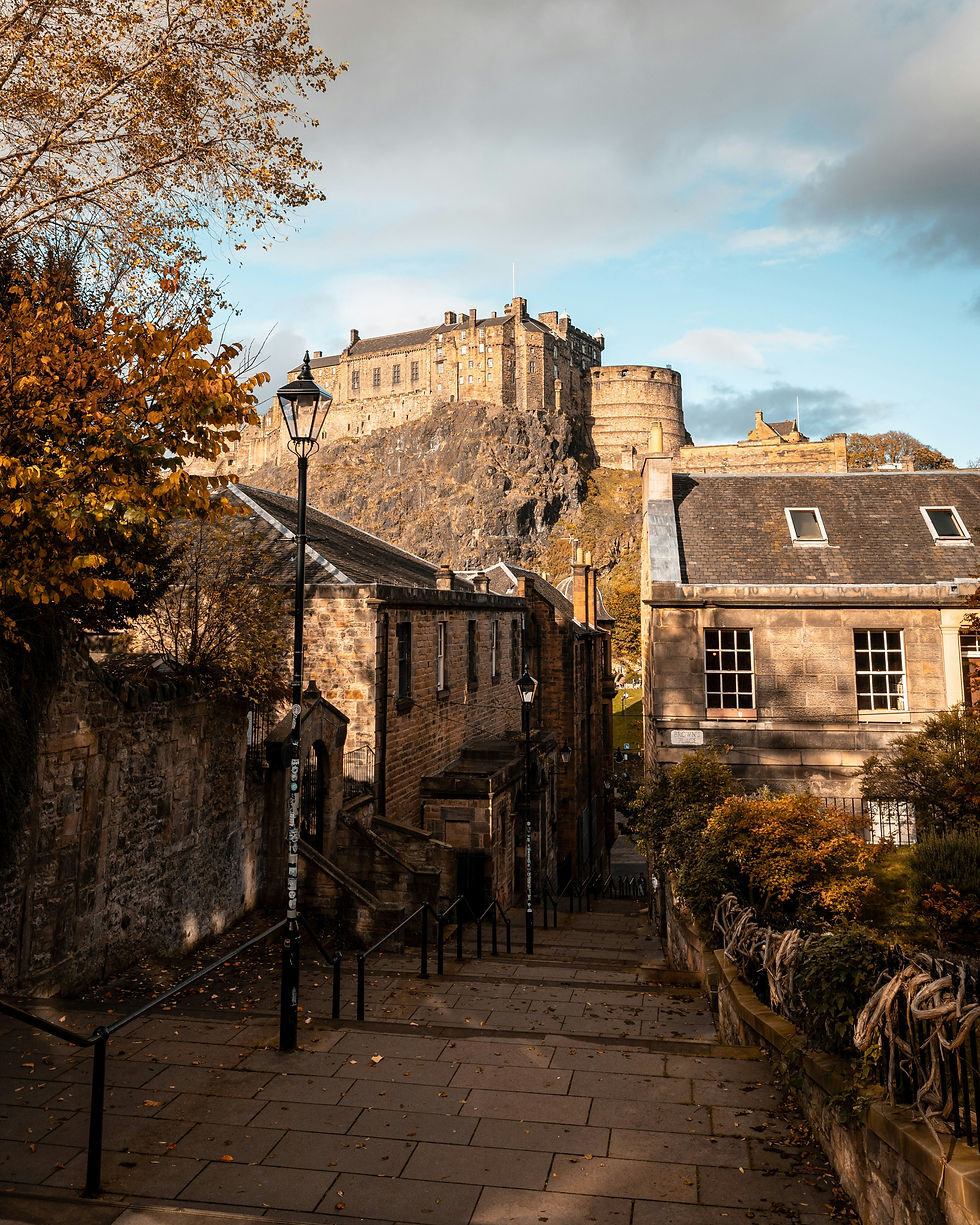 Edinburgh Castle as viewed from the Vennel Steps with the roofs of houses in the foreground, a lamppost, and a tree with orange leaves.