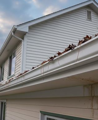 Clogged white seamless gutters filled with dry leaves and pine needles on a two-story home, showing water runoff stains on the beige siding.