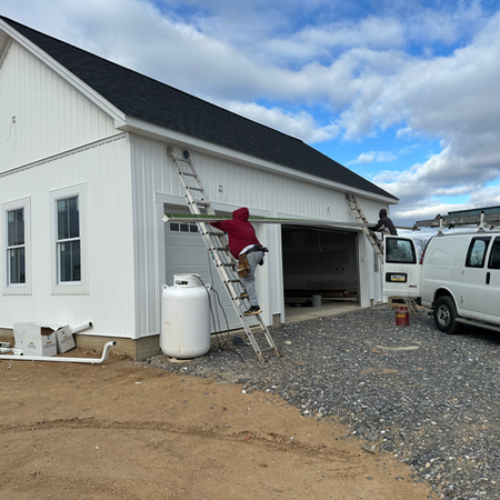 Construction progress on attached modern farmhouse garage in Westminster, MD: workers on ladders installing seamless gutters by Shepherd & Sons Inc., white board-and-batten siding, black roof, triple garage doors, under partly cloudy blue sky with work van and materials on-site.