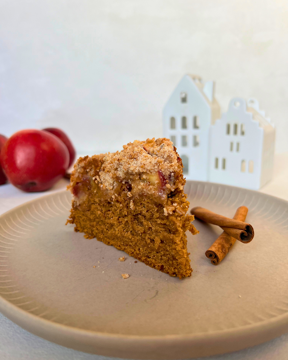 Ein Stück Apfelkuchen, 3 rote Äpfel, 2 Zimtstangen, beige Teller, heller Untergrund