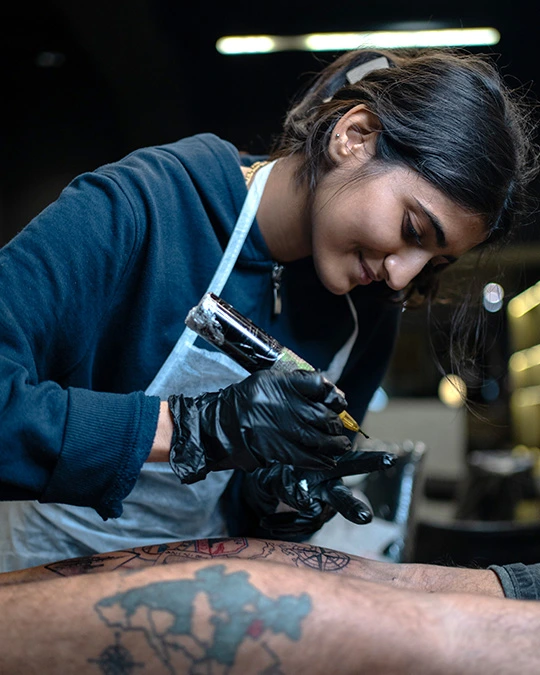Grishita Suthar tattoo artist at Aliens tattoo working on a client's leg, detailing a travel-themed tattoo with intricate map and symbols.