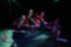 Five people in black with red scarves perform with brooms onstage, intense expressions, colorful lighting, seated man watches.