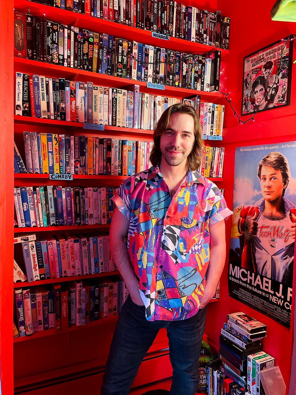Man in a colorful shirt stands in a vibrant red room filled with VHS tapes. A "Teen Wolf" poster and string lights are visible in the background.