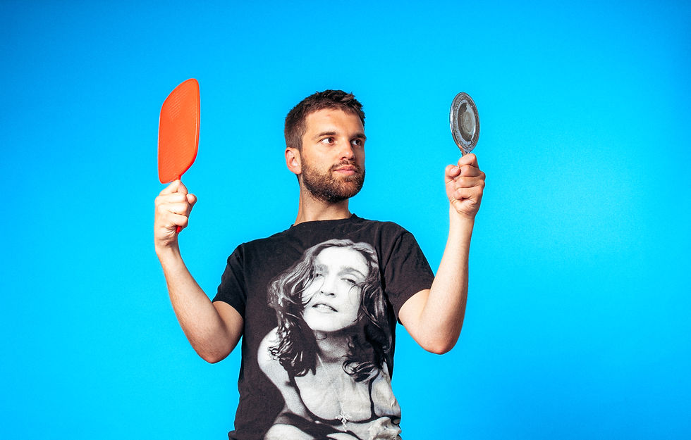 Will Owen holding two mirrors and wearing a Madonna T-shirt, standing in front of a bright blue background with a playful expression.