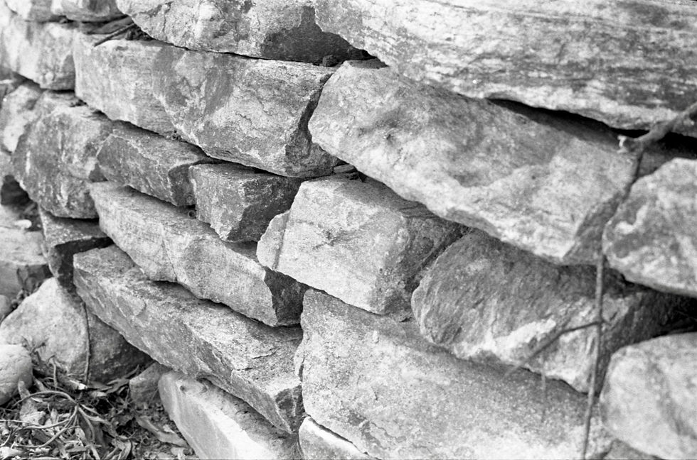 Black-and-white close-up of a stacked stone wall, with rough blocks and a few leaves and twigs at the base.