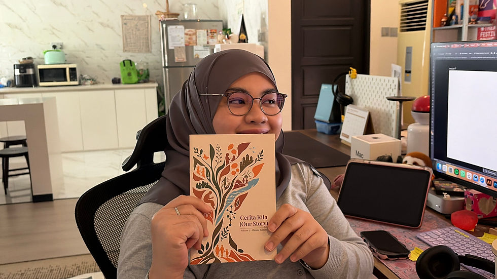 Majidah Khirul in hijab and glasses holds up Cerita Kita (Our Story) in a home office with a computer and kitchen behind.