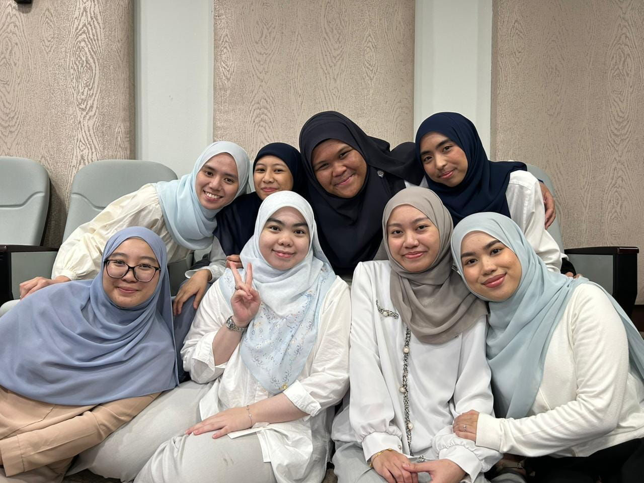 A group of eight women in hijab smiling and posing, some doing a peace sign, wearing pastel and neutral colours  