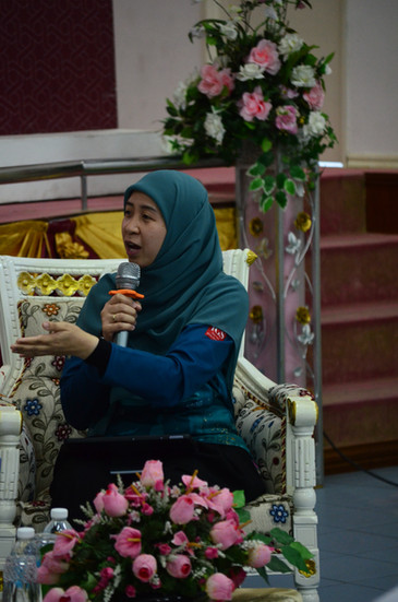 A lady in emerald hijab and blue shirt talking into the microphone, sat on a floral couch