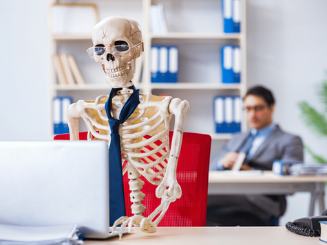 A skeleton, wearing a tie, sat behind a laptop at a desk, in an office environment