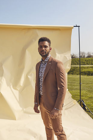 Uomo in completo marrone caramello monopetto abbinato a camicia floreale bianca e blu, con spilla decorativa sul bavero. 