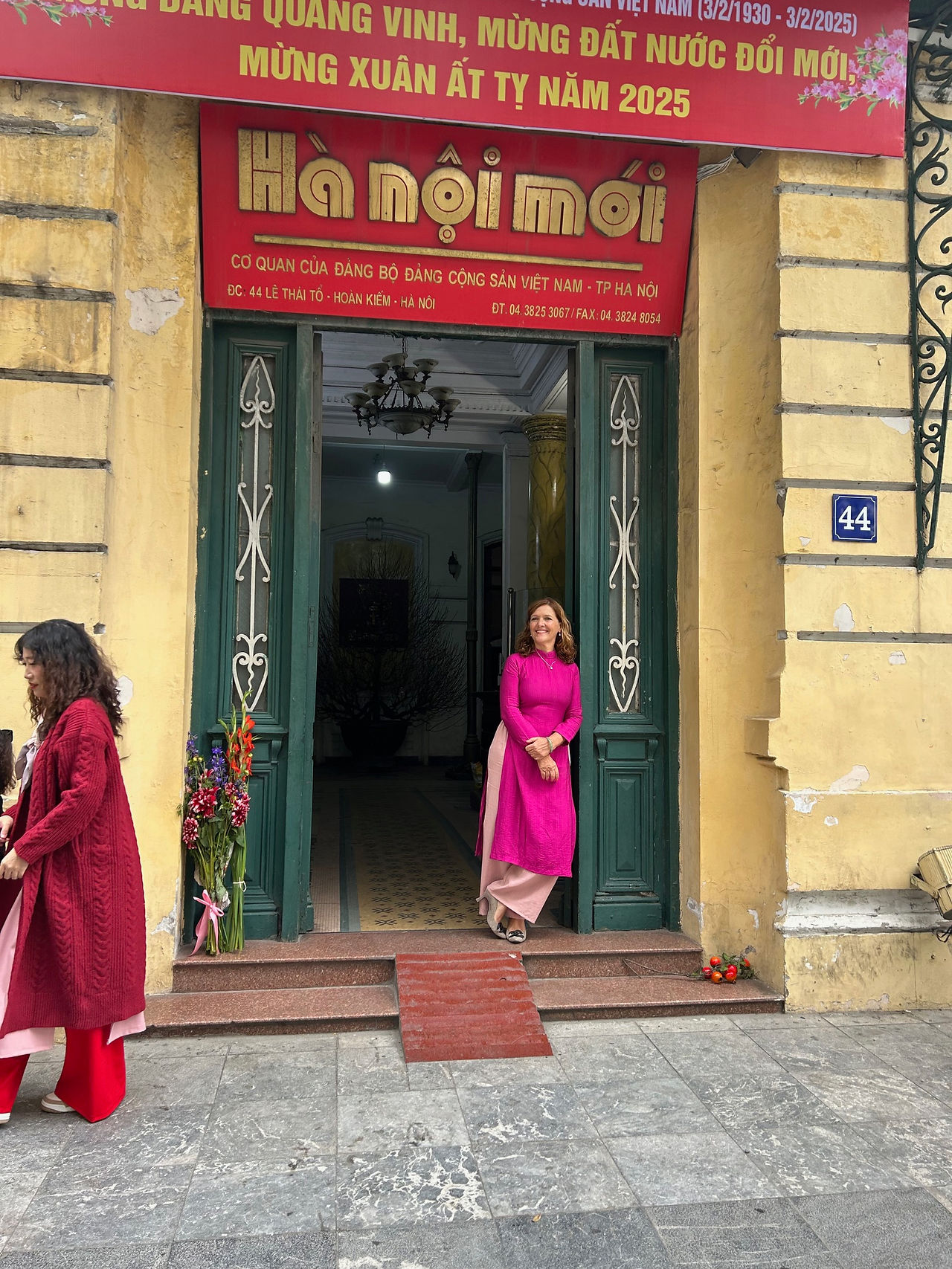 A woman in a red traditional outfit leans against a dark green wooden doorframe of a vintage-style building. A red sign with yellow lettering hangs above the entrance, and the architecture features classic textured walls and a small wooden ramp at the doorstep.