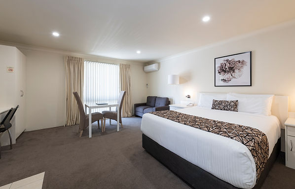 Executive King Room with large king bed with white linens behind there is a table for two a sofa a bright window and an air con and cabinet on the edge