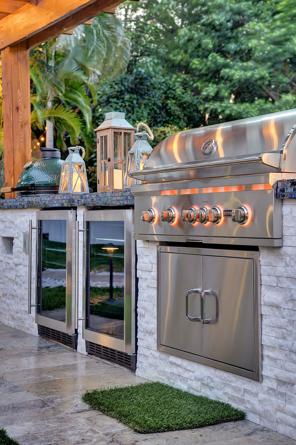 Outdoor kitchen with stainless steel grill, stone tiles, and lanterns on countertop. Lush greenery in background. Cozy and inviting mood.
