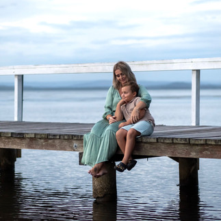 A tender scene of mum and her young son sitting side by side on the edje of the jetty, cuddling and chatting softly as they look out over the water.