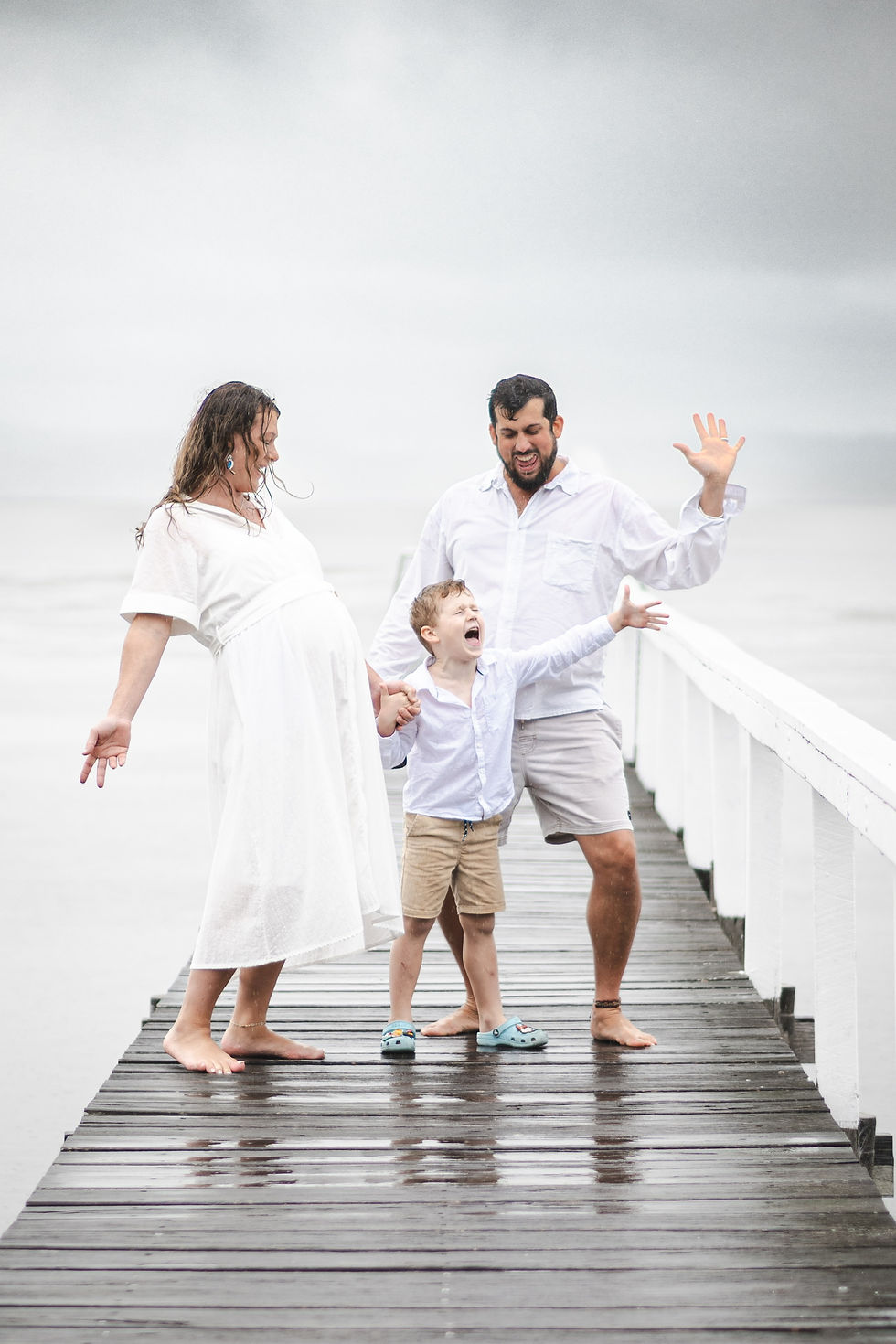 A family of three expecting twins, stands together on the Long Jetty pier in the rain, joyfully yelling “pizza”!