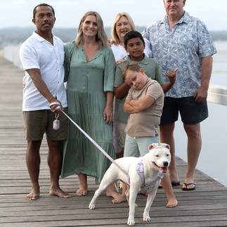 A family including mum, dad, their children, her parents and their dog, pose on together in coordinating green and white outfits at Long Jetty, smiling warmly in the soft afternoon light.