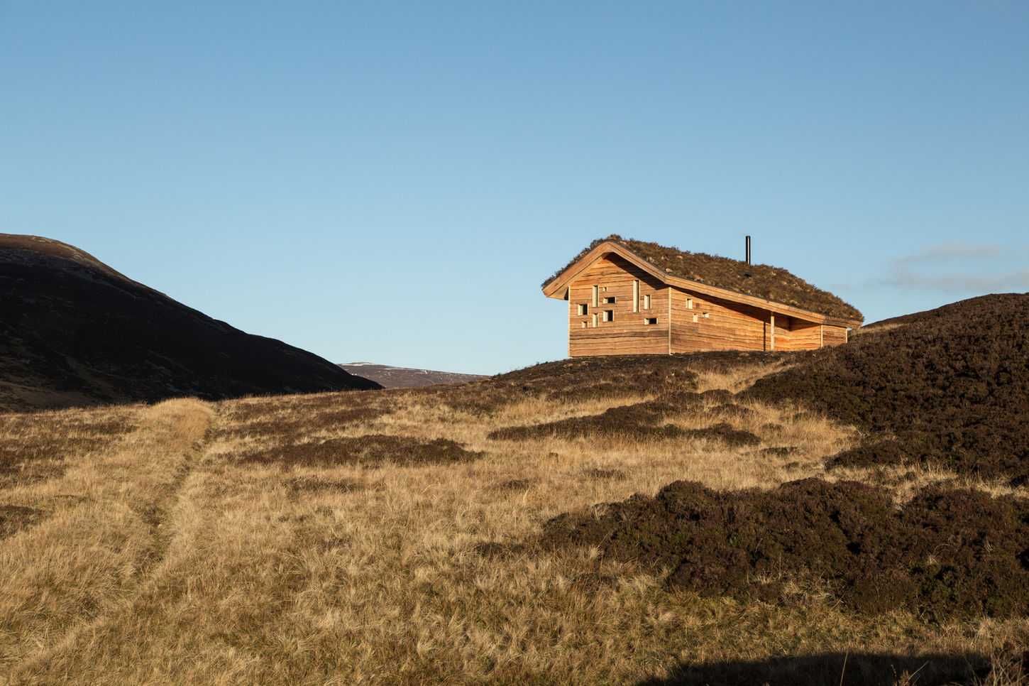 "Tucked away in a remote and rugged part of the Cairngorms National Park in the Scottish Highlands, the Culardoch Shieling is a small mountain hut designed by London and Aberdeenshire-based firm Moxon Architects. The 47-square-meter hut is a contemporary reinterpretation of traditional Scottish farming crofts, shepherd's shelters, and Swiss Alpine shacks. It was commissioned by art dealers Iwan and Manuela Wirth, who sought a private, remote space on their estate for entertaining guests.
The design is a clever blend of regional vernacular architecture and 20th-century modernism. Its monolithic, wooden form draws inspiration from the simple aesthetic of local livestock buildings, while its irregular, strategically placed windows are reminiscent of Le Corbusier's iconic Chapelle Notre Dame du Haut at Ronchamp. These irregularly cut apertures precisely frame specific features of the surrounding landscape, including the granite tors of Ben Avon and a bend in the River Gairn.
Constructed almost entirely from timber, the shieling (Gaelic for ""hut"") was designed to minimize its impact on the terrain. The structure features a simple cruck frame with deep, overhanging eaves. The roof is camouflaged with a layer of heather, moss, and stone gathered from the local hillsides, helping it blend into the moorland and providing additional insulation.
The cozy interior is lined with spruce and features a long, communal dining table, a prominent wood-burning stove for heating, and subtle modern details like a suspended iron shelf for candles. The project was completed in 2016 and received several awards, including the Royal Incorporation of Architects in Scotland Forestry Commission for Scotland Award in 2017.
Project credits
Architects: Moxon Architects
Photography: Ben Addy
Main Contractor: AJC Aboyne
Project Manager: Fowler Fortescue
Structural Engineer: Graeme Craig Consulting Engineers Ltd
Quantity Surveyor: McCue & Porter
Manufacturers: Velux Group, Glenalmond Timber, Tor Contracting"