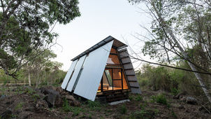 Named after the suliformes genus of seabirds, the SULA cabin by architect Diana Salvador is a prefabricated, A-frame home designed for the unique environment of the Galápagos Islands. Completed in 2023 on Santa Cruz Island for a local family, the project showcases an innovative approach to sustainable construction by minimizing environmental impact in a delicate ecological region.
The two-story, modular cabin was prefabricated over two months in Quito, Ecuador, before its 2,000 custom-made components of wood, metal, aluminum, and glass were shipped in containers to the island. The components, held together by over 17,000 screws and pins, were assembled on-site in just four weeks by a specialized team. This prefabrication process avoided construction site damage to the soil.
Inspired by bioclimatic principles, the cabin was elevated off the ground to allow for a cooling air chamber beneath it. A double-layered wall and roof structure, along with perforated floors, promote natural cross-ventilation, ensuring a comfortable interior climate. The five main materials—wood, stone, metal, glass, and PVC—were chosen for their efficiency and minimal ecological footprint. The interior features plywood paneling, creating a warm, inviting atmosphere that contrasts with the exterior.
The main A-frame structure creates a double-height central space. A loft accessed by a sliding black metal ladder offers a flexible area for sleeping, working, or leisure. Large windows at each end of the cabin, along with two narrow windows on the sides, fill the space with natural light and frame views of the surrounding nature.
The cabin was intentionally designed with scalability and relocation in mind, using gabions as foundations to further minimize the impact on the soil.
Credits
Architects: Diana Salvador
Client: Catalina and family
Prefabrication: Huaira
Photography: JAG Studio
Construction: Lahden Puurakentajat
Transport: 2 trucks, 2 containers