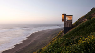 Located in Matanzas, Chile, the cabins La Loica and La Tagua were designed by Croxatto y Opazo Arquitectos as minimalist holiday homes that blend with the surrounding coastal environment. The project is defined by its strategic position on a steep, wind-swept hill, offering expansive views of the Pacific Ocean.
The project's design focused on creating small-footprint volumes that respect the natural landscape while maximizing coastal views. Named after native bird species—La Loica and La Tagua—the cabins use vertically stacked rooms to create a lookout-tower effect.
The cabins are situated on Lagunilla hill in Matanzas, Navidad, approximately 80 meters above sea level and 2.5 hours from Santiago. From this vantage point, they overlook the "Lobera," a large rock formation in the
The cabins are built entirely of wood, using a timber pylon structure to support the main platform on the steep terrain. The exterior cladding is made from reclaimed oak sleepers treated with petroleum oils to protect against marine corrosion. Interiors feature a limited palette of light-colored wood, creating a warm, simple, and spacious atmosphere.
The living area, kitchen, and bathroom are located on the main floor, with a ladder leading to the bedroom in the loft. Large, west-facing windows enhance the feeling of hovering over the ocean and dominate the horizon. North-facing terraces extend the interior spaces and are positioned to provide a shield against the region's strong southwest winds.
To combat the small footprint (La Loica at 20 m² and La Tagua at 25 m²), the double-height living room creates a perception of more expansive space.
The project initially operated off-grid, requiring the use of a water tank and electric generator for the construction process. The cabins were designed for two different clients, and the firm’s main goal was to connect the inhabitants with nature.
Project credits
Architects: Croxatto y Opazo Arquitectos.
Lead Architects: Nicolás Opazo Marchetti and Felipe Croxatto Viviani.
Photography: Cristóbal Palma / Estudio Palma.