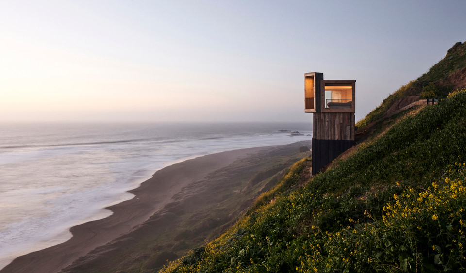 Located in Matanzas, Chile, the cabins La Loica and La Tagua were designed by Croxatto y Opazo Arquitectos as minimalist holiday homes that blend with the surrounding coastal environment. The project is defined by its strategic position on a steep, wind-swept hill, offering expansive views of the Pacific Ocean. 

The project's design focused on creating small-footprint volumes that respect the natural landscape while maximizing coastal views. Named after native bird species—La Loica and La Tagua—the cabins use vertically stacked rooms to create a lookout-tower effect.
The cabins are situated on Lagunilla hill in Matanzas, Navidad, approximately 80 meters above sea level and 2.5 hours from Santiago. From this vantage point, they overlook the "Lobera," a large rock formation in the 

The cabins are built entirely of wood, using a timber pylon structure to support the main platform on the steep terrain. The exterior cladding is made from reclaimed oak sleepers treated with petroleum oils to protect against marine corrosion. Interiors feature a limited palette of light-colored wood, creating a warm, simple, and spacious atmosphere.

The living area, kitchen, and bathroom are located on the main floor, with a ladder leading to the bedroom in the loft. Large, west-facing windows enhance the feeling of hovering over the ocean and dominate the horizon. North-facing terraces extend the interior spaces and are positioned to provide a shield against the region's strong southwest winds.

To combat the small footprint (La Loica at 20 m² and La Tagua at 25 m²), the double-height living room creates a perception of more expansive space.

The project initially operated off-grid, requiring the use of a water tank and electric generator for the construction process. The cabins were designed for two different clients, and the firm’s main goal was to connect the inhabitants with nature. 

Project credits
Architects: Croxatto y Opazo Arquitectos.
Lead Architects: Nicolás Opazo Marchetti and Felipe Croxatto Viviani.
Photography: Cristóbal Palma / Estudio Palma.