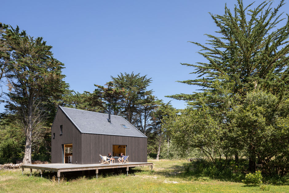 Le Refuge KE01 is a black timber cabin nestled in the dunes of Keremma in Brittany, France, designed by Gayet Roger Architects as a family vacation retreat. Built in 2019, the 850-square-foot cabin (79 square meters) is positioned on a raised platform supported by four-foot-tall metal stilts, which protects it from the area's risk of flooding and preserves the natural terrain. A wooden footbridge connects the entrance of the house to a small path that leads to the nearby beach.
The cabin's design draws inspiration from traditional Scandinavian architecture, featuring a dark exterior that allows it to blend into the surrounding landscape of tall cypress trees. The cross-laminated timber (CLT) structure is insulated with blown-in cellulose wadding and clad with rough-sawn wood strips painted with black Falun-style paint, a durable Swedish pigment. At night, the house disappears into the darkness, with only the illuminated windows visible like hanging lanterns.
Inside, the warm, spruce-clad living spaces are designed with function in mind, stripped of the superfluous. The architects used rough-sawn wood panels for the built-in benches, dining table, shelving, and counters. The open-plan space is punctuated by strategically placed windows that frame specific views of the landscape. The large fixed window in the corner seating nook offers pastoral scenery, while a western bay window can be opened to extend the main room onto the terrace, blurring the line between inside and outside.
Upstairs, the four sleeping areas are designed like small cabins. Each features small, eye-level windows that provide views of the landscape for those lying down and can accommodate two single beds, with a built-in bench on one side and a storage area on the other.
Credits
Architects: Gayet Roger Architects (Aldric Gayet & Anne Gayet)
Location: Tréflez, Finistère, France
Completion Year: 2019
Photography: Pascal Leopold
Foundation: Techno Pieux Nantes Nord
Wood Structure, Insulation, Exterior Joinery, Partitioning & Joinery: SARL Construibois
Plumbing, Sanitary, Ventilation, Electricity & Wood Stove: SARL Kerbaul
Roofing: SARL BCI
