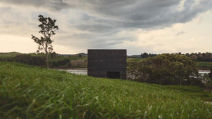 Completed in 2014, the Eyrie Cabins are a pair of small, autonomous holiday homes designed by New Zealand-based Cheshire Architects. Sited on a grassy hill overlooking the Kaiwaka Harbour, near the Tasman Sea, the cabins were conceived as a polemic against the trend of overly large and ostentatious holiday homes. Each cabin measures just 29 square meters, embracing a minimalist philosophy to intensify the connection between occupant and nature. 

The design is a bold response to its isolated, pristine setting, with the two cabins deliberately positioned low on the slope rather than on the hilltop plot that was originally assigned. This decision was made to make the structures less conspicuous and to help them feel like an intimate part of the landscape rather than something that dominates it. 

The cabins are clad in burnt black timber, a treatment inspired by the abstract painter Kazimir Malevich. This creates a stark, monolithic form that contrasts dramatically with the vivid green of the hillside and helps the structures blend into the landscape.
In a radical departure from traditional cabin design, the Eyrie Cabins have no doors. Entry is gained by climbing a series of boulders and entering through a pull-down window. This subversion of standard building language was intended to make the small structures feel like something more than just houses—like small, autonomous vessels.
Large, oblong windows are strategically placed to frame specific views of the coast and the hillside, while solid side walls ensure privacy between the two cabins. This limited fenestration creates a deeply interior, contemplative experience, contrasting with the typical desire for "indoor-outdoor flow" in coastal architecture.
The cabins embody a philosophical stance against architectural decadence. By using minimal materials and a compact form, the project advocates for a more thoughtful and humble vision for coastal architecture in New Zealand. 

While the cabins present a stark, matching exterior, their interiors are beautifully and uniquely individualized for the two separate clients. The interior design emphasizes luxury through thoughtful detailing rather than opulent size. 
Cabin 1: Features a small, brass-clad kitchen, adding a jewel-like touch of luxury to the dark space.
Cabin 2: Showcases the rich grain of oiled jarrah wood, which contrasts with the lighter tones of standard construction plywood.
The material palette is restrained but carefully selected to enhance the serene and introspective atmosphere. 

Project credits
Architect: Cheshire Architects (led by Nat Cheshire and Ian Scott).
Photography: Jeremy Toth.
