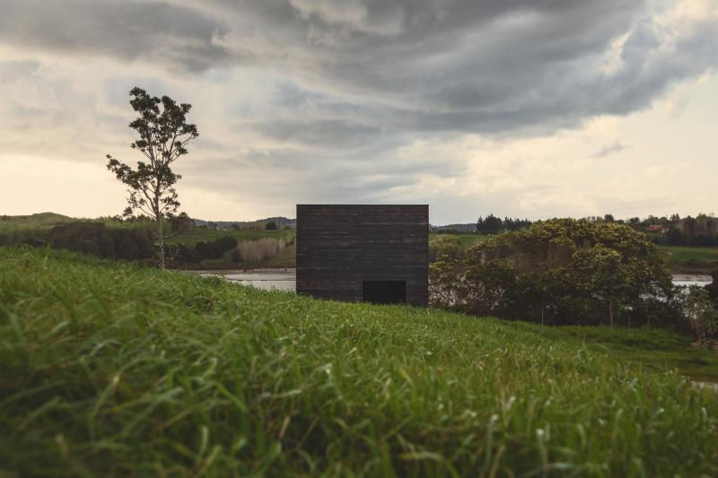 Completed in 2014, the Eyrie Cabins are a pair of small, autonomous holiday homes designed by New Zealand-based Cheshire Architects. Sited on a grassy hill overlooking the Kaiwaka Harbour, near the Tasman Sea, the cabins were conceived as a polemic against the trend of overly large and ostentatious holiday homes. Each cabin measures just 29 square meters, embracing a minimalist philosophy to intensify the connection between occupant and nature.
The design is a bold response to its isolated, pristine setting, with the two cabins deliberately positioned low on the slope rather than on the hilltop plot that was originally assigned. This decision was made to make the structures less conspicuous and to help them feel like an intimate part of the landscape rather than something that dominates it.
The cabins are clad in burnt black timber, a treatment inspired by the abstract painter Kazimir Malevich. This creates a stark, monolithic form that contrasts dramatically with the vivid green of the hillside and helps the structures blend into the landscape.
In a radical departure from traditional cabin design, the Eyrie Cabins have no doors. Entry is gained by climbing a series of boulders and entering through a pull-down window. This subversion of standard building language was intended to make the small structures feel like something more than just houses—like small, autonomous vessels.
Large, oblong windows are strategically placed to frame specific views of the coast and the hillside, while solid side walls ensure privacy between the two cabins. This limited fenestration creates a deeply interior, contemplative experience, contrasting with the typical desire for "indoor-outdoor flow" in coastal architecture.
The cabins embody a philosophical stance against architectural decadence. By using minimal materials and a compact form, the project advocates for a more thoughtful and humble vision for coastal architecture in New Zealand.
While the cabins present a stark, matching exterior, their interiors are beautifully and uniquely individualized for the two separate clients. The interior design emphasizes luxury through thoughtful detailing rather than opulent size.
Cabin 1: Features a small, brass-clad kitchen, adding a jewel-like touch of luxury to the dark space.
Cabin 2: Showcases the rich grain of oiled jarrah wood, which contrasts with the lighter tones of standard construction plywood.
The material palette is restrained but carefully selected to enhance the serene and introspective atmosphere.
Project credits
Architect: Cheshire Architects (led by Nat Cheshire and Ian Scott).
Photography: Jeremy Toth.