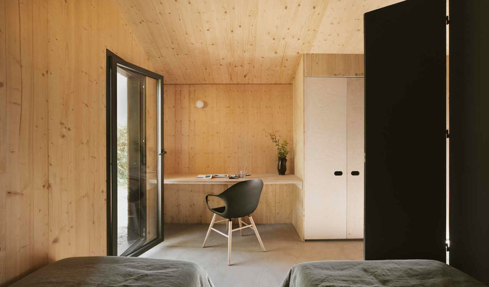 Pine Nut Cabane, designed by Daab Design, is a timber-clad retreat set within a pine forest and olive grove near Marseille, France. Created for a multi-generational family, the cabin serves as a guest studio, artist's workspace, and rural escape. Its design prioritizes flexibility and a deep connection to its natural surroundings.
The minimalist design, with its simple volume and use of local materials, references traditional regional farming architecture. The cabin is intended as a subtle, understated space that enhances the client's favorite spot on their property, a place for painting and playing pétanque.
The 35 m² cabin is carefully positioned on a rural property outside Marseille, hidden from both the main farmhouse and local roads. It is oriented to the east to capture the gentle morning sunlight and offer expansive valley views. A dense pine forest on the south side provides natural shading from the intense Mediterranean sun.
The cabin is clad in scorched pine, a durable, locally sourced material that allows the structure to blend with the forest landscape. The interior features a combination of light plywood panels, matte-black surfaces, and polished concrete floors. This provides a tranquil, balanced atmosphere that contrasts with the bright outdoor light.
The design incorporates deep eaves, high-spec double glazing, and cross-ventilation to minimize heat gain and keep the interior cool naturally. The polished concrete floor is integral to the thermal heating and cooling strategy.
The cabin's open-plan layout can be reconfigured with full-height matte-black doors to create a central corridor or two private bedrooms. A central, matte-black pod contains the bathroom, which features a cave-like shower with terracotta zellige tiles. An east-facing terrace with a fire pit provides an outdoor seating area with panoramic views. Built-in storage and desks ensure the space remains uncluttered and functional for various activities like yoga, painting, or sleeping.
Project credits
Architects: Daab Design.
Director: Anaïs Bléhaut.
Builder/Contractor: Moustache Bois.
Landscape Design: Tamaris Design.
Photography: Henry Woide.
Masonry/Ground Works: Joos.
Glazing: Technal.
Tiles: Art & Sud Marseille.
Interior Furniture: Vitra.
Exterior Furniture: Fermob.