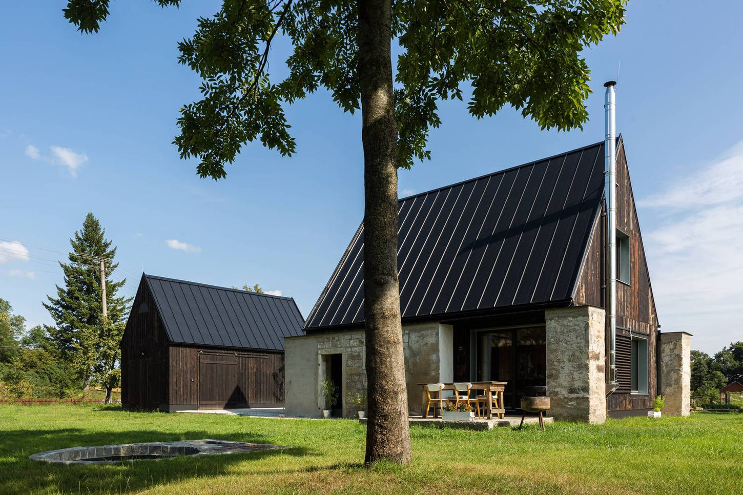 "House for Parents in Jeseniky is a family residence designed by TRI.ŠTRNÁST architekti and completed around 2021 in the Czech Republic. Situated near the Jeseníky Mountains, the project involved transforming a plot where a barn once stood. Instead of demolishing the original structure entirely, the architects salvaged the monumental granite walls, inserting a new timber construction between them to preserve the site's history. The new house is connected to a separate studio/garage via a central, multifunctional outdoor terrace.
The design was created for an elderly couple who wished to move from a city apartment to a house with a garden. The primary living space is on the ground floor and is designed to accommodate the couple's future needs, featuring a no-threshold layout and a bathroom accessible to wheelchairs. This main floor includes an entry foyer, bathroom, utility room, living room with dining area and kitchen, and a bedroom. A staircase leads to an attic that serves as a bedroom for visiting family and provides storage for sports equipment for the nearby Ramzová ski area.
The house emphasizes sustainability and low-energy performance. The KVH (solid constructional wood) lumber beam system was assembled with minimal fasteners and insulated with blown cellulose. For durability and aesthetic continuity with local architecture, the exterior is clad in charred spruce wood, an application of the shou sugi ban technique. The interior features perimeter walls lined with unfired bricks and finished with clay plaster, which helps regulate humidity and creates a comfortable indoor climate. Heating is provided by a solid fuel stove, with electric cables as a backup. The southern facade is complemented by a stone terrace shaded by a textile canopy, preventing the building from overheating.
Credits
Architects: TRI.ŠTRNÁST architekti
Photography: Lukáš Ildža
"