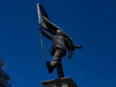 A gray statue proudly holds a flag that is suffocating its face, continuing to walk forward even though they're about to walk off the ledge holding them up.