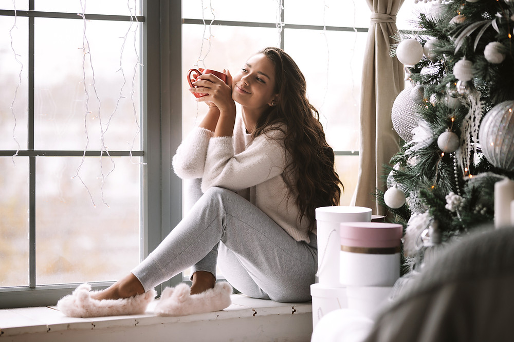Smiling woman with long brown hair, dressed in lounge wear and fuzzy slippers sitting on window seat next to christmas tree and holding a cup of hot chocolate.