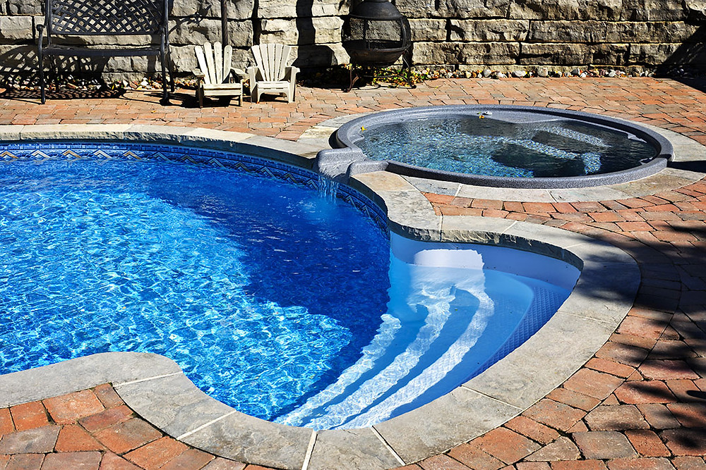 Round pool with steps, brick patio, stone wall in background, and hot tub flowing into pool.