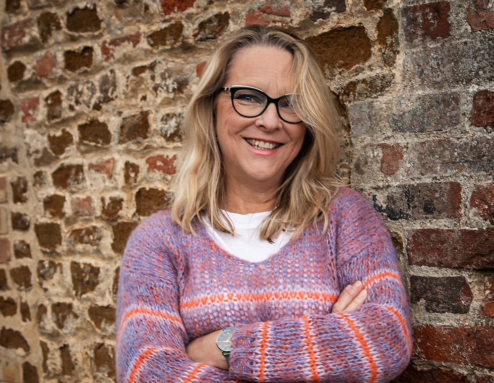 Smiling Sarah J Naylor, arms crossed, brick wall background