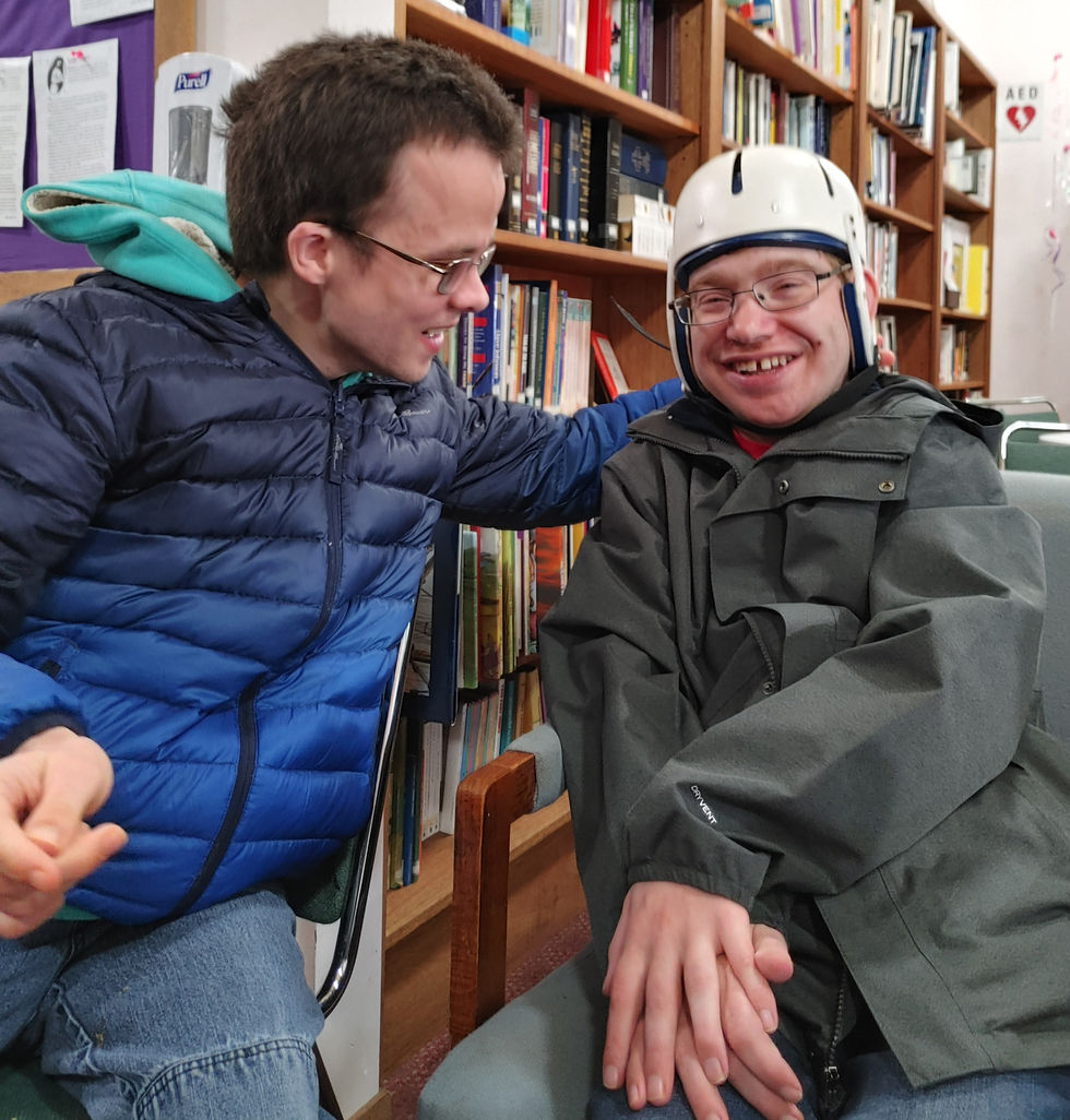 One young man puts a welcoming arm around another young man, each of them is sitting down in a chair and they're both smiling