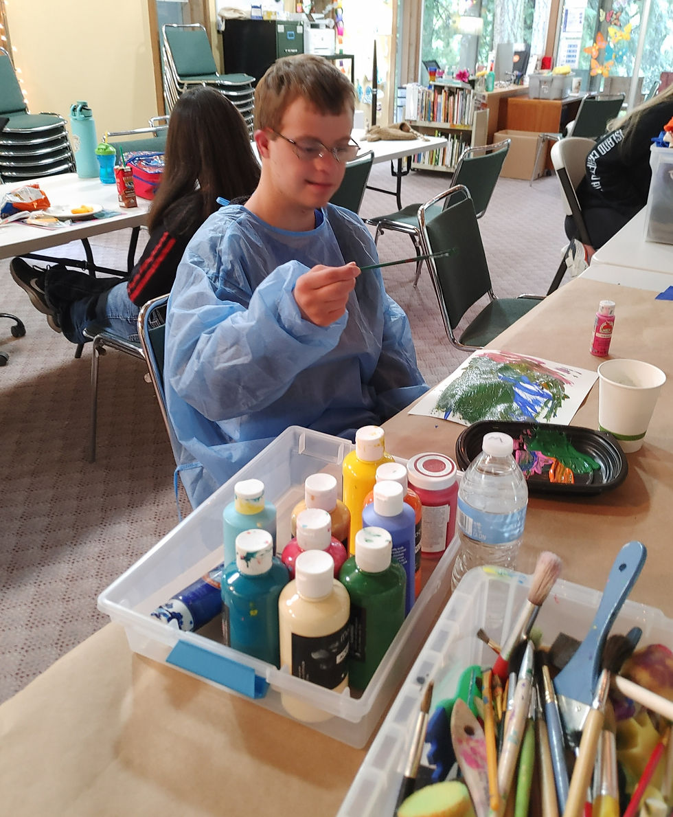 A young man holds a paintbrush above his artwork. He's sitting at a table with lots of arts supplies, while a friend eats lunch at a different table behind him.
