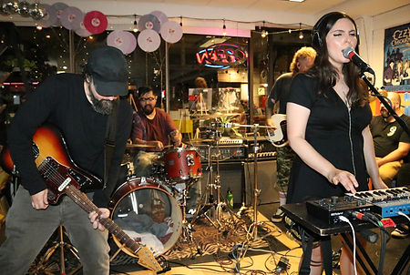 A photo of One Hundred Moons performing at Kops records. In the foreground is a guitarist and a lead singer on keys. in the back is a drummer and a bassist. 