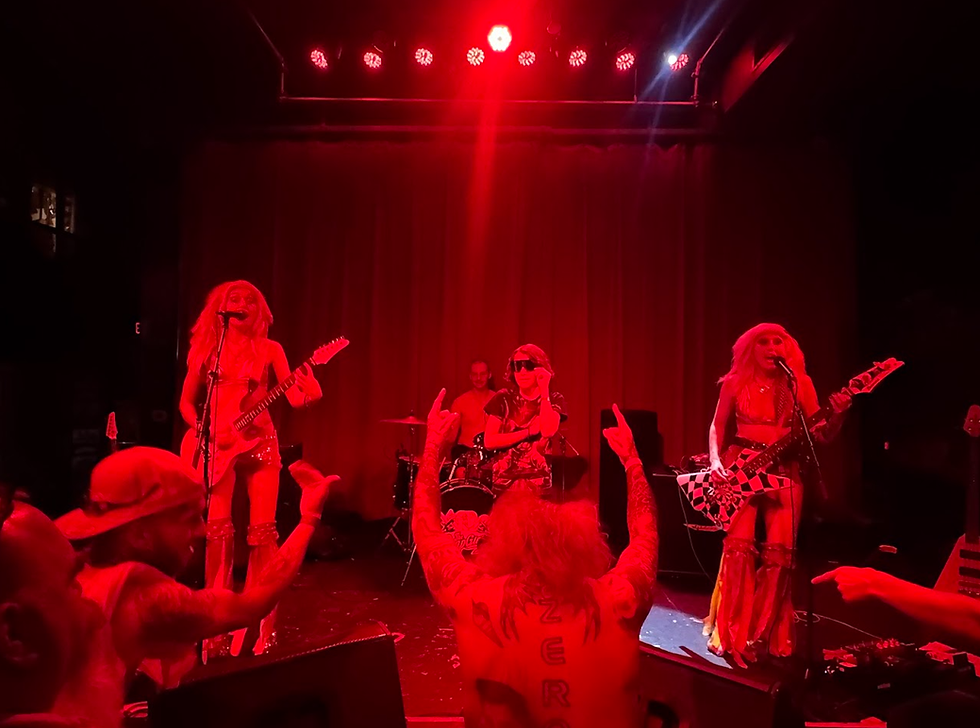 All-female punk rock band performs on stage under intense red lighting, with two guitarists and a drummer commanding the spotlight while an energetic, tattooed crowd throws up rock hand signs. The performers wear bold, glittery outfits and long blonde wigs, creating a glam-meets-grunge atmosphere at this high-octane live show.